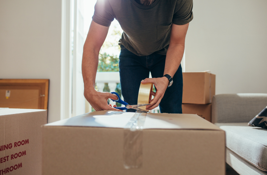 A man packing cardboard boxes with tape and he is cutting the tape.