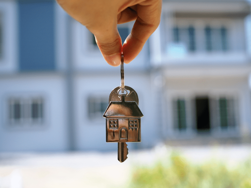 Person holding their house key in front of their new home.