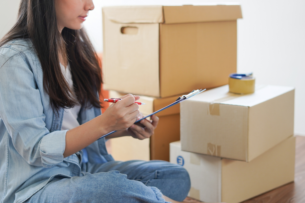 Woman sitting on the floor next to cardboard boxes checking off items on a clipboard.