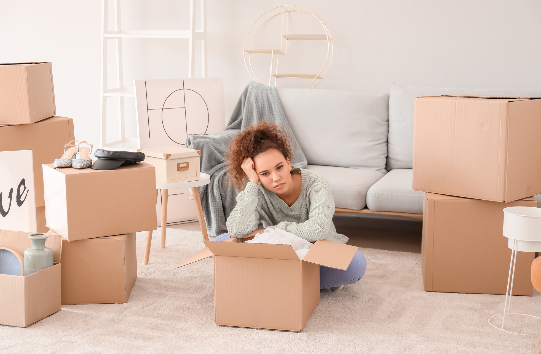 Woman sitting on the floor, frustrated while leaning on an open cardboard box, surrounded by other cardboard boxes.