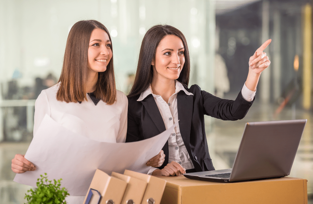 Two women smiling standing in front a laptop and holding open a blueprint while one points.