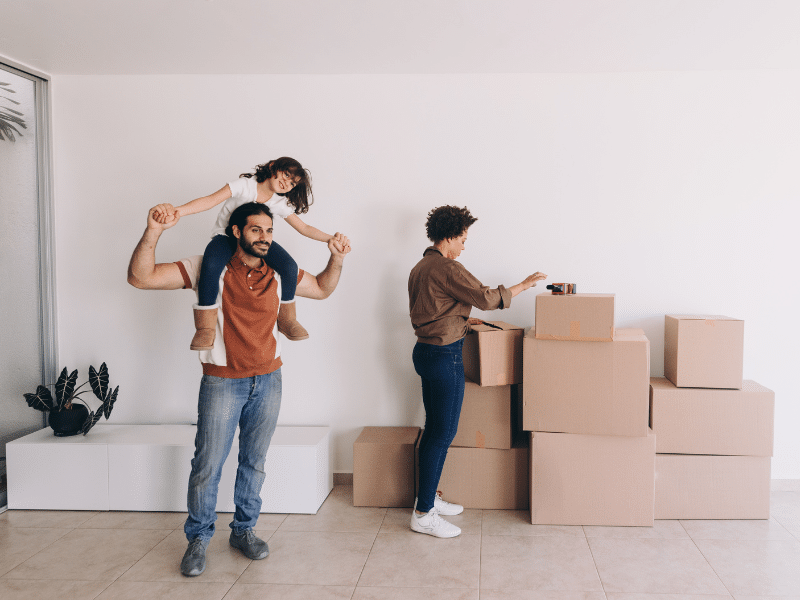 Dad with daughter on his shoulders standing next to his wife while she is going through cardboard moving boxes.