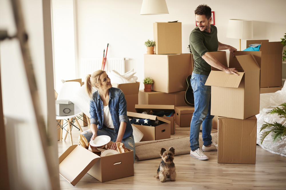 Man smiling looking back at a woman sitting on the ground with their dog in between them while they both unpack cardboard boxes.