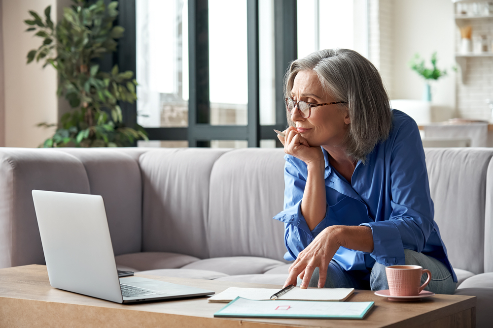 Older woman looking a computer while holding a pen sitting on a couch.