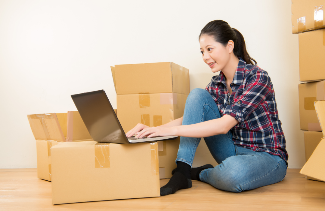 A woman looking at a laptop surrounded by cardboard boxes.