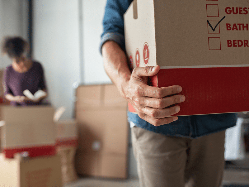 Man carrying a cardboard box with a woman reading a book in the background.