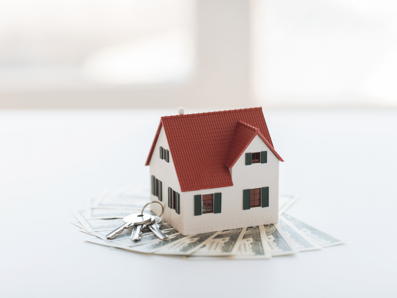 Small wooden house with house keys sitting on dollar bills.