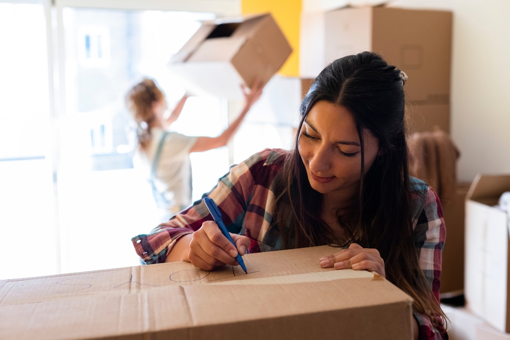 A woman writing on a box with another woman moving a box behind her.