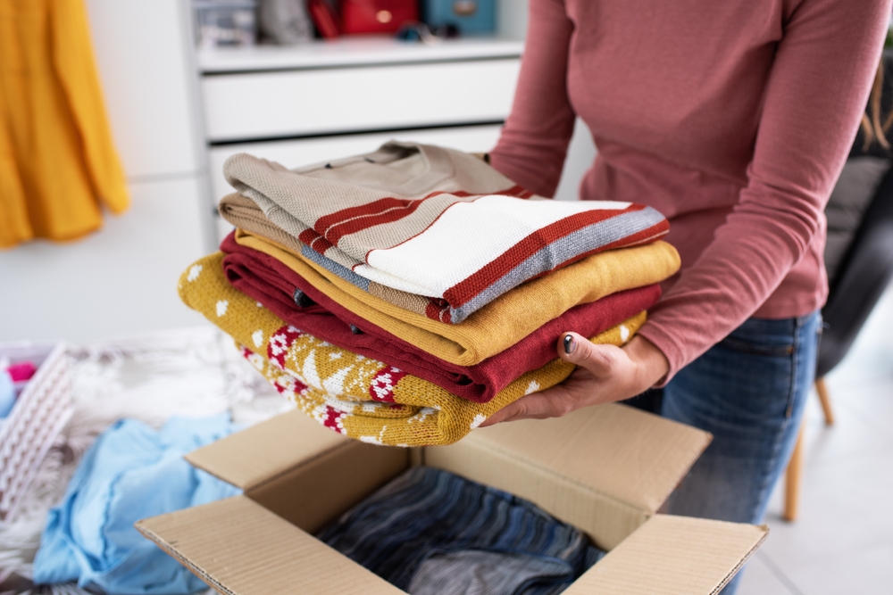 A woman putting folded clothes into a box surrounded by more clothes.