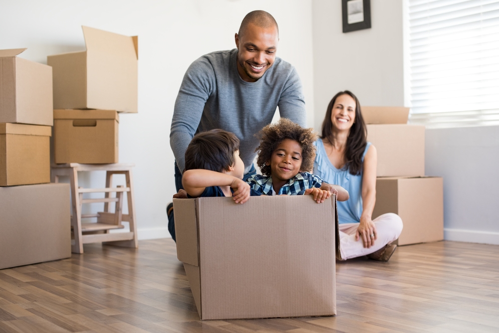 Man pushing his kids in a cardboard box across the floor while his wife smiles behind him.