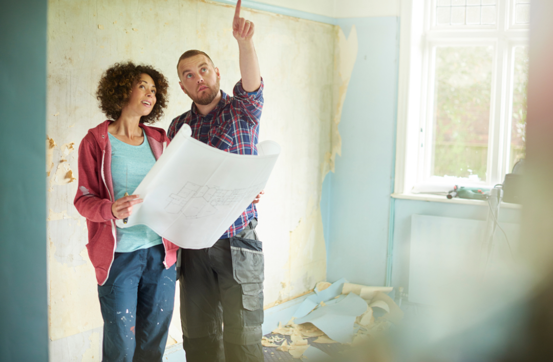 Woman holding blueprints while a man points at the ceiling in a room mid repair.