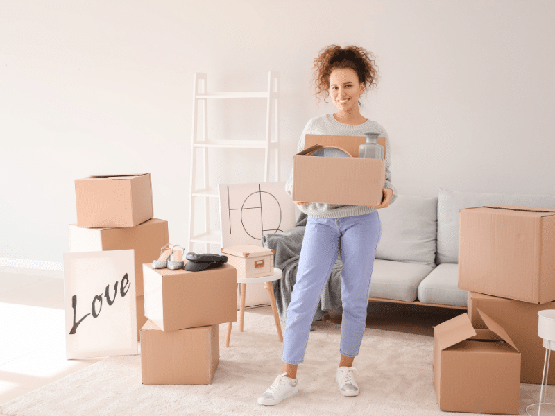 Woman standing in a packed up living room holding a cardboard moving box.