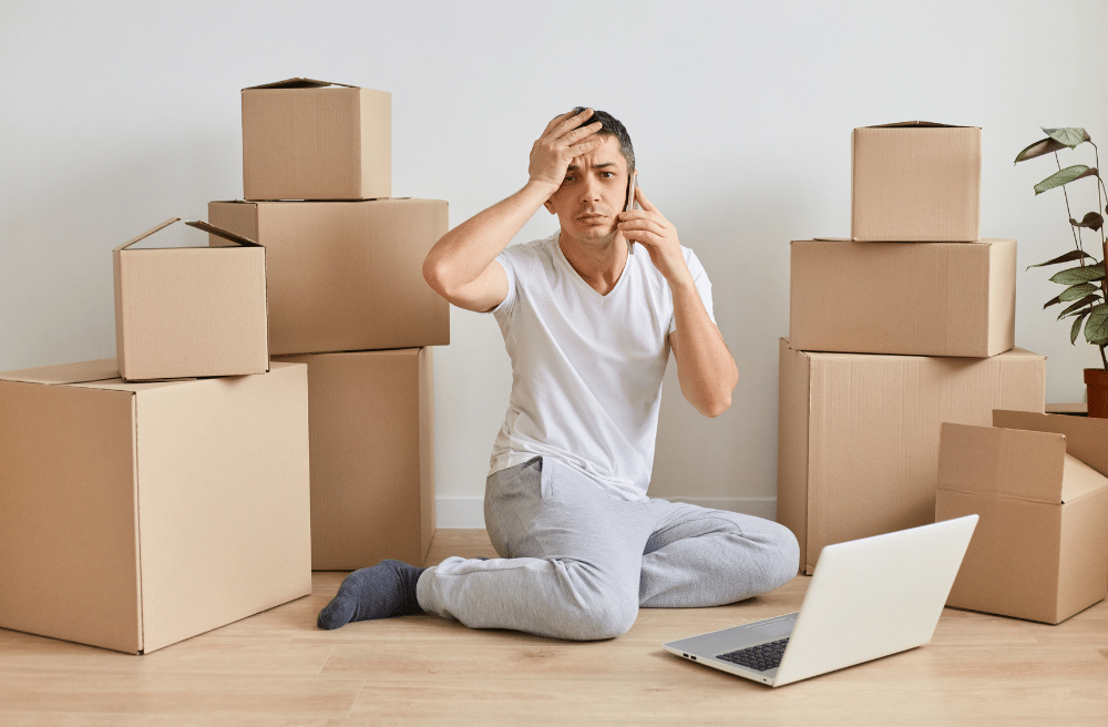 A Man sitting on the ground with his laptop looking stressed surrounded by boxes.