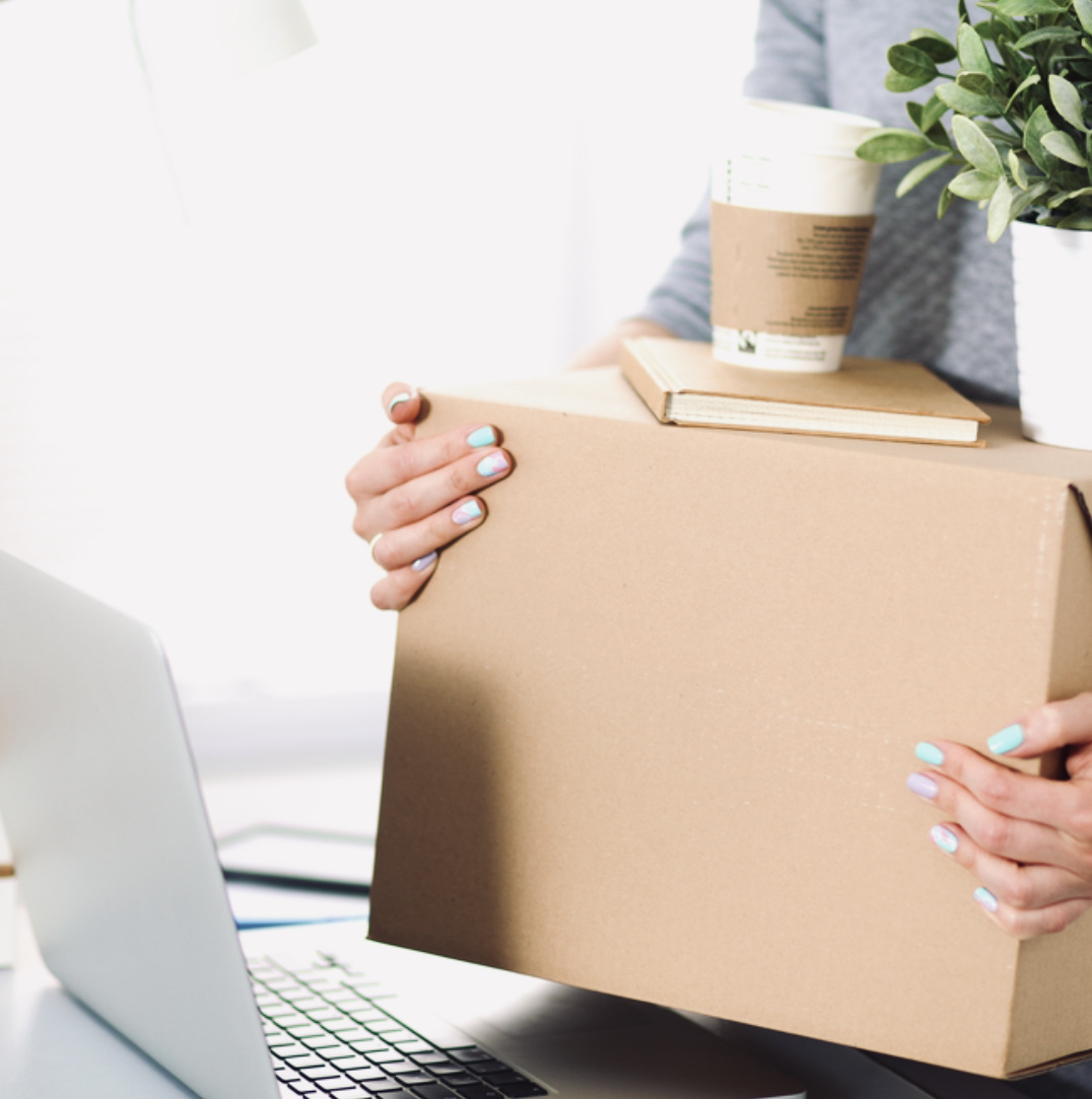 Woman carrying a cardboard box with a small book, coffee cup and potted plant on top of it.