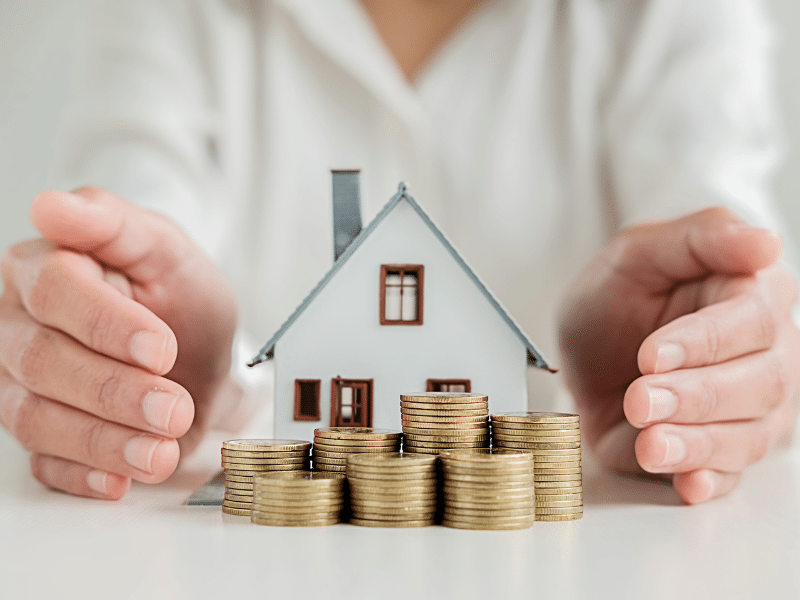 Hands cupping a miniature house with coins in front of the house.