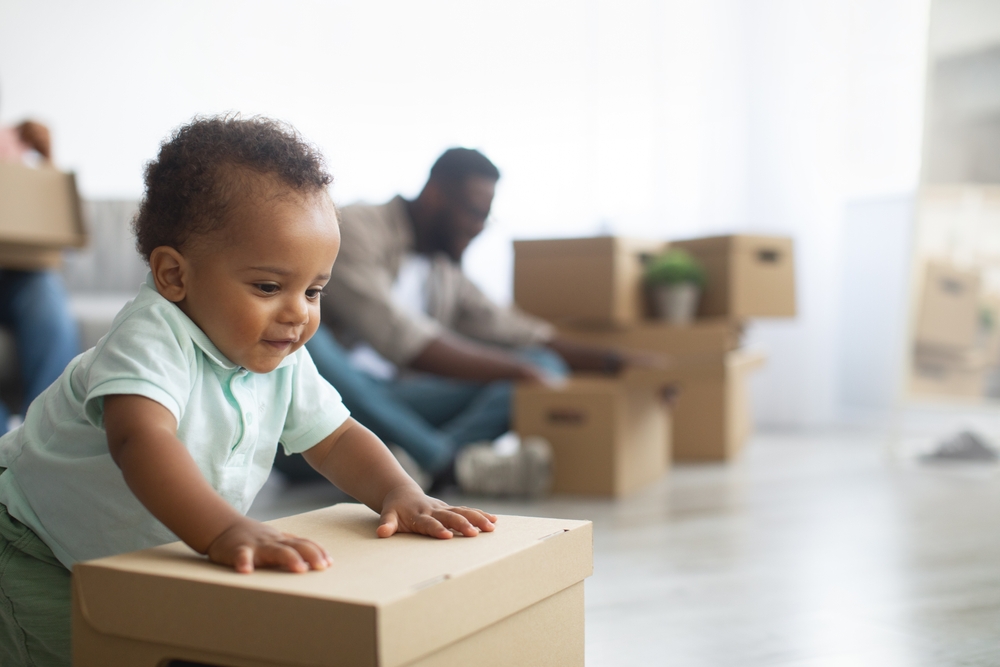 Baby pushing a cardboard box lid closed while his parents sit behind him.