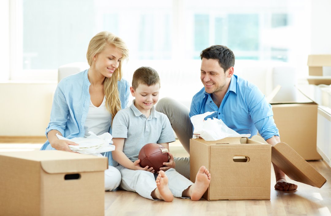 Parents sitting on the floor with their son holding a football surrounded by cardboard boxes.