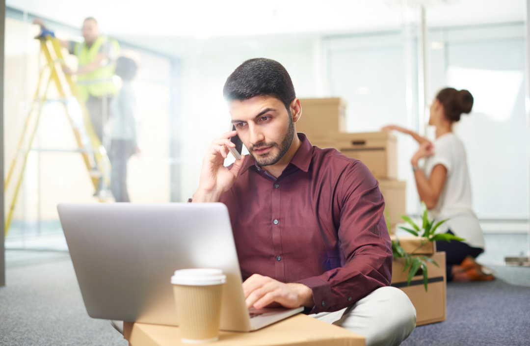 Man sitting on the floor working on his laptop while his partner sits behind him and people work on their house.