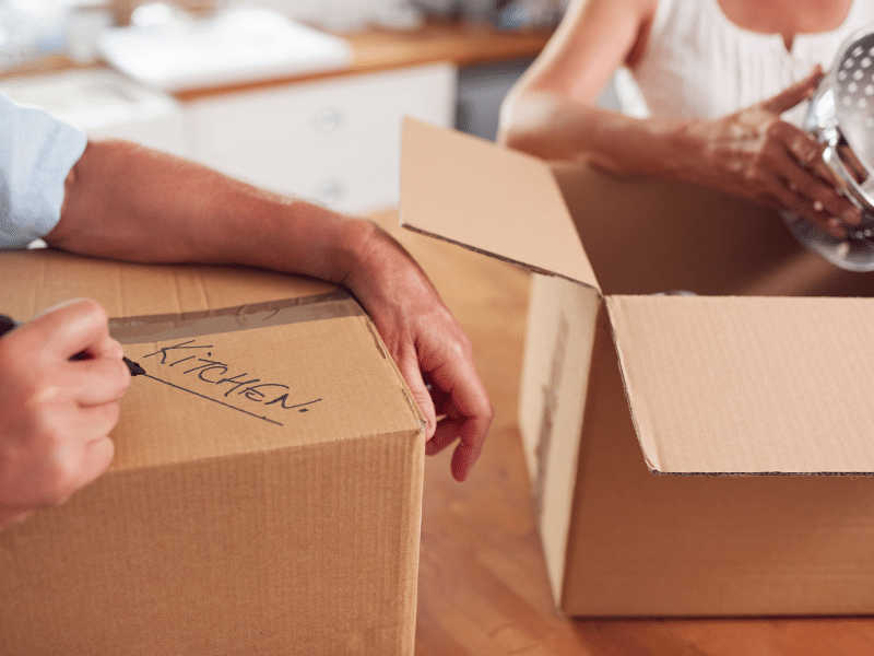 Couple packing up cardboard boxes and labeling them kitchen.