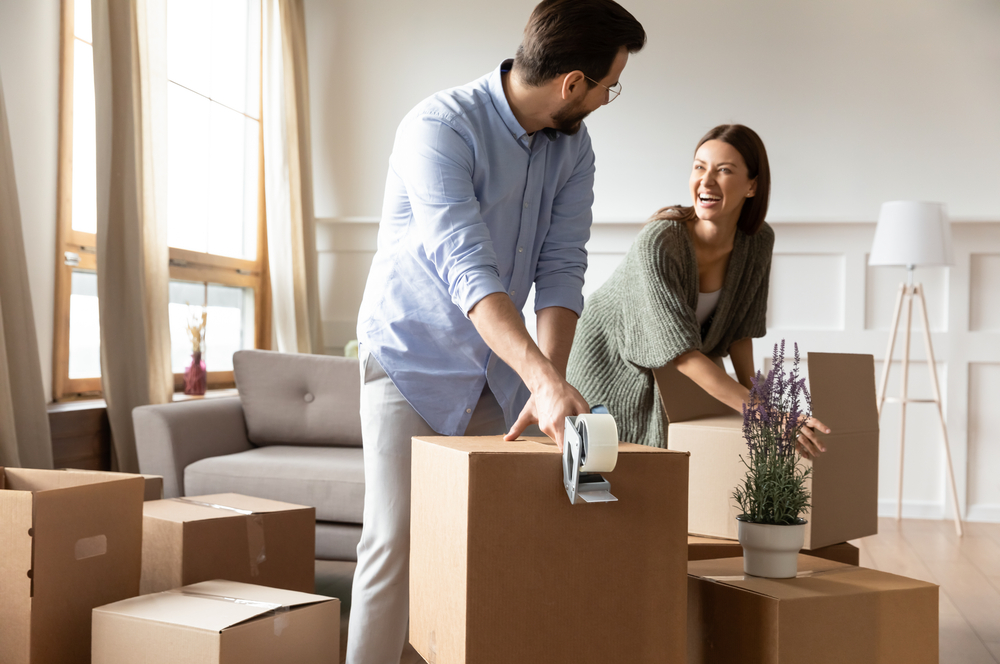 Couple smiling at each other while taping cardboard boxes closed.