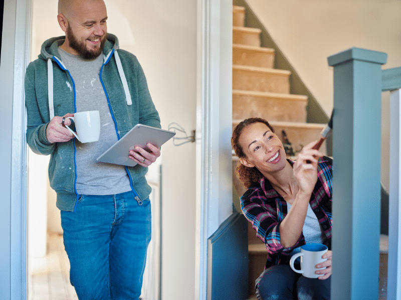 Man carrying a cup of coffee and an IPad leaning against a doorframe watching his wife paint their stair case.