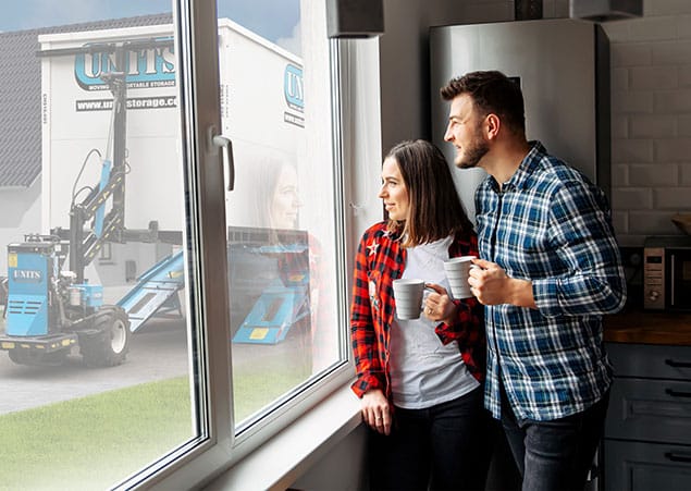 couple looking out the window while drinking coffee. Outside of the window is a UNITS moving and portable storage container is getting dropped off